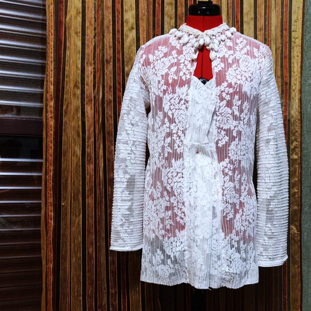 Long sleeve woman blouse in white pleated lace with floral patterns. The geometric neckline is embellished with raw linen trim and natural white seashell pendants. Fastened with a seashell button featuring a golden rim.