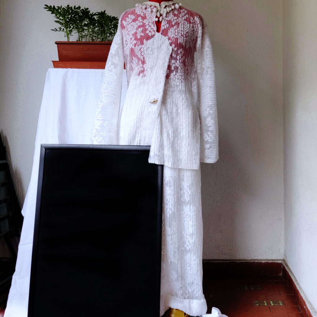 Woman set in white floral macramé, long sleeved blouse and widetrousers. The blouse features a geometric neckline adorned with raw linen trim and natural white seashell pendants. Fastened with a golden seashell button. The pants have a cuffed hem and inner cotton lining that subtly reveals the legs. This style is a perfect option for a seaside wedding.