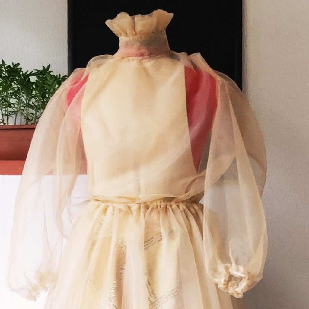 Long-sleeved lady blouse in transparent beige silk chiffon. The high ruffled collar is enhanced by tone-on-tone embroidery and fastened at the back with raw pearl buttons. Poet style sleeves with elastic cuffs.