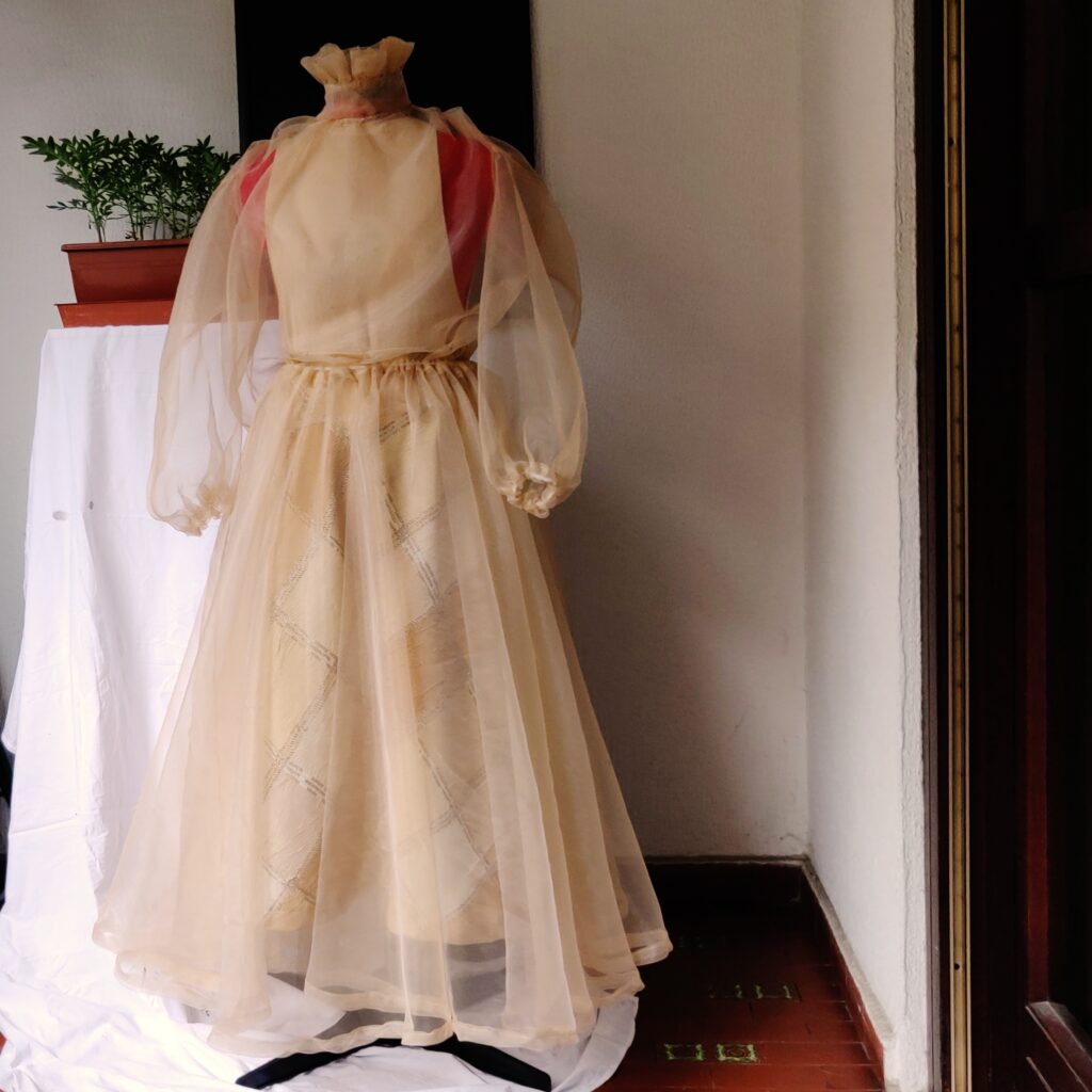Woman set in transparent beige silk chiffon of a long-sleeved blouse and mid skirt. The blouse has a high ruffled collar with embroidery and fastens at the back with pearl buttons. Poet style sleeves with elastic cuffs. The full skirt is wide, flowing, and ultra-light, with an elastic waistband. This look is an elegant solution for a beach wedding or any other special occasion.